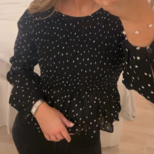 Svart blus med vita prickar - Säljer en söt svart blus med vita prickar och volangdetaljer. Blusen har långa ärmar med knappar vid ärmsluten och en smickrande passform. Perfekt för att piffa upp vardagslooken eller för en kväll ute! 💗