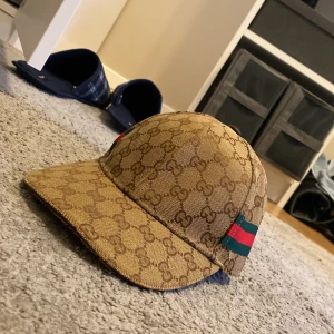 Beige keps från Gucci - Snygg beige keps från Gucci med det klassiska GG-mönstret. Kepsen har en justerbar rem i brunt skinn och detaljer i grönt och rött på sidorna. Perfekt för att ge din outfit en lyxig touch.