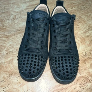 Loubs  - Snygga svarta sneakers med nitar på tån och sidorna. Skorna har snörning och en röd sula som ger en cool kontrast. Perfekta för att ge din outfit en edgy touch.