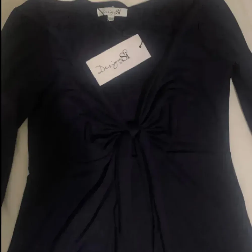 Snygg svart långärmad topp från Design by si med knytning framtill. Perfekt för en stilren look med en touch av elegans. Toppen har en djup v-ringning och är tillverkad i ett mjukt material.. Puserot.