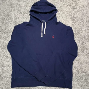 Blå hoodie från Polo Ralph Lauren - Säljer en snygg blå hoodie från Polo Ralph Lauren med en klassisk röd logga på bröstet. Hoodien har en justerbar huva med vita snören och en praktisk magficka. Perfekt för en avslappnad stil.