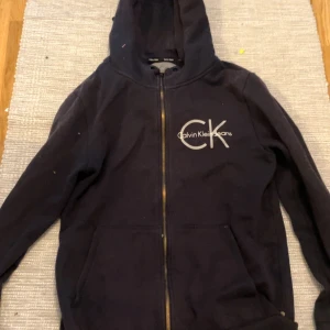 Intressekoll 02 - Säljer en svart hoodie från Calvin Klein med dragkedja och stor CK-logga på bröstet. Den har en bekväm passform och är perfekt för en avslappnad stil. Huvan ger extra värme och stil. Perfekt för vardagsbruk!