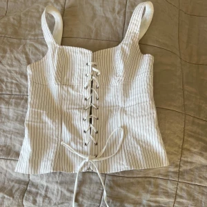 Vit randig korsettopp från Zara - Säljer en snygg vit korsettopp från Zara med smala bruna ränder och snörning. Dock en liten rosa röd fläck på insidan men syns absolut inte på utsidan. Perfekt för en stilren look!