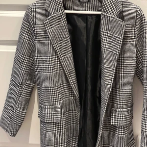 Rutig kappa från Boohoo - Snygg rutig kappa från Boohoo i svart och vit. Den har en klassisk krage och långa ärmar. Perfekt för vår med sitt stilrena mönster och bekväma passform.