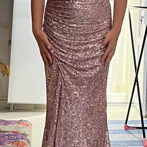 Glittrig rosa långklänning - Säljer en fantastisk glittrig rosa långklänning med draperad design. Klänningen har en vacker omlott-look och är perfekt för festliga tillfällen. Den är axelbandslös och har en elegant passform som framhäver figuren.