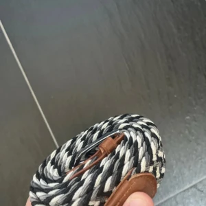 Randigt bälte med läderdetaljer - Snyggt randigt bälte i svart och grått med bruna läderdetaljer. Perfekt för att ge en stilren touch till din outfit. Bältet har ett unikt mönster och är lätt att justera.