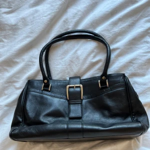 Svart handväska från House of Couture - Elegant svart handväska i skinn från House of Couture. Väskan har dubbla handtag och en dekorativ spänndetalj framtill. Den är utrustad med dragkedja för säker förvaring av dina tillhörigheter. Perfekt för en stilren look. Så himla snygg! Priset kan diskuteras!!❤️💞