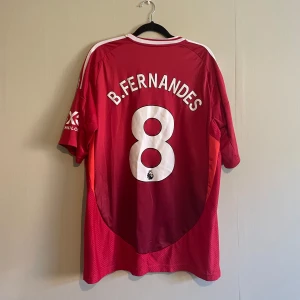  Manchester United fotbollströja 24/25 - (Det står XL i den men den passar för L)Säljer en röd Manchester United fotbollströja med B. Fernandes och nummer 8 på ryggen. Tröjan har korta ärmar och Premier League-logga på ärmen. Den är prydd med klubbens emblem på bröstet och har vita detaljer på axlarna.