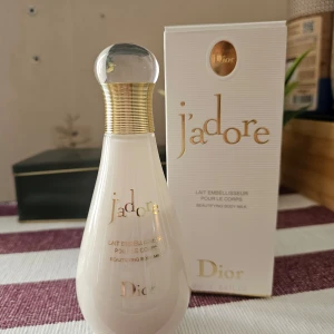 J'adore Beautifying Body Milk från Dior - Elegant kroppsmjölk från Dior i serien J'adore. Flaskan har en stilren design med en mjuk, pärlemorskimrande yta och en gyllene kork. Förpackningen är vit med gyllene detaljer, vilket ger en lyxig känsla. Perfekt för att återfukta och ge huden en subtil doft.