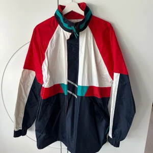 Flerfärgad regnjacka med retrostil - Säljer en cool flerfärgad regnjacka i retrostil med färgerna röd, vit, svart och turkos. Jackan har en hög krage och dragkedja framtill. Perfekt för en sportig look eller en promenad i stan en regnig dag. Har lite fläckar 
