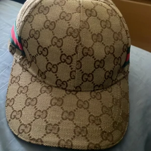 Beige keps från Gucci - Snygg beige keps från Gucci med det ikoniska GG-mönstret. Kepsen har gröna och röda detaljer på sidan och är tillverkad i Italien. Perfekt för att ge din outfit en lyxig touch.
