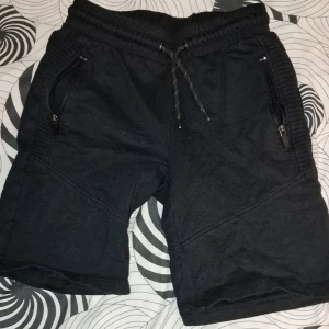 Svarta shorts från Core med dragkedjor - Svarta shorts från Core med elastisk midja och snörning. Shortsen har två fickor med dragkedja framtill och är tillverkade i ett mjukt material. Perfekta för vardagsbruk eller träning.