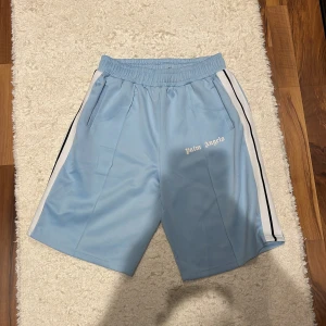 Ljusblå shorts från Palm Angels med vita och svarta ränder - Snygga ljusblå shorts från Palm Angels med vita och svarta ränder längs sidorna. Shortsen har elastisk midja och sidofickor. Perfekta för en avslappnad stil.