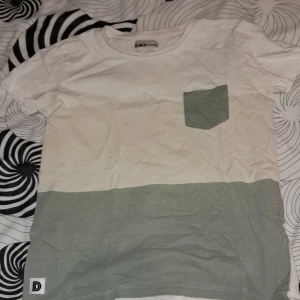 Flerfärgad t-shirt med bröstficka från D.M.D - Säljer en t-shirt från D.M.D i tre färgblock: vit upptill, beige i mitten och ljusgrön nedtill. T-shirten har en matchande ljusgrön bröstficka och korta ärmar. Perfekt för en avslappnad stil.