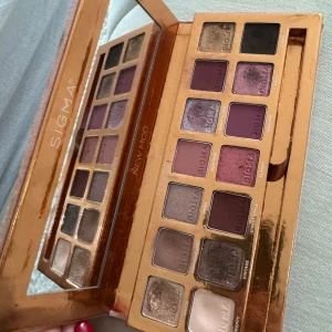 SIGMA ögonskuggspalett med spegel - En lyxig ögonskuggspalett från SIGMA med 14 nyanser i både matta och skimrande toner. Färgerna sträcker sig från neutrala beige och bruna till lila, guld, svart och vinrött. Paletten har en stor spegel i locket och guldfärgad ram.