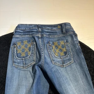 Blå bootcut jeans med broderade bakfickor - Snygga blå bootcut jeans från Violet med guldiga broderier på bakfickorna. Klassisk femficksmodell med låg midja och guldfärgade detaljer vid knappar och nitar. Perfekta för dig som gillar en unik touch på dina jeans.