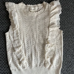 Vit stickad ärmlös topp med volang  - Supersöt vit stickad topp med spetsvolanger längs axlarna. Ärmlös modell med fint mönsterstickat tyg och rund halsringning. Perfekt för sommarens outfits!