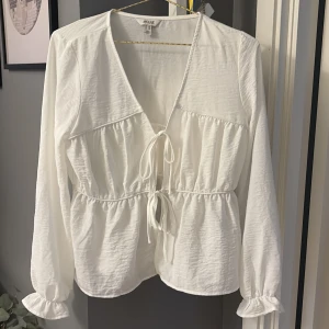 Vit blus med knytning - Säljer en vit blus från AWARE med v-ringning och knytning framtill. Blusen har långa ärmar med resår vid ärmslut och en lätt rynkad midja för en feminin siluett. Perfekt till vardags och till lite mer festligare. 
