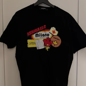 Svart t-shirt från Dsquared2 med Milano-tryck - Säljer en svart t-shirt från Dsquared2 i storlek XL men passar Medium & Large. Framsidan har färgglada tryck med texten 'Milano Dreamin', Dsquared2-logga och olika patch-inspirerade motiv. Perfekt för dig som gillar streetwear och statement-plagg. 