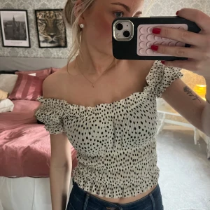 Prickig offshoulder topp från NA-KD - Supersöt offshoulder topp från NA-KD i vitt med svarta prickar. Smockad och croppad modell med volangdetaljer vid ärmar och halsringning. Perfekt för vår och sommar, lätt att matcha med jeans eller kjol.