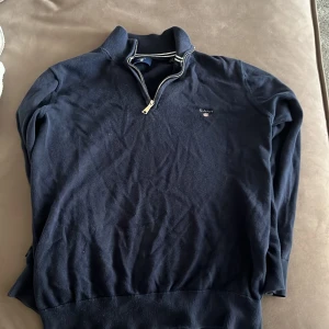 Mörkblå halvzip tröja från Gant - Säljer en mörkblå tröja från Gant med halv dragkedja och ståkrage. Tröjan har ribbade muddar och en broderad Gant-logga på bröstet. Perfekt för en stilren och avslappnad look.