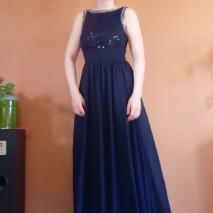 Mörkblå balklänning med paljetter - Mörkblå balklänning med paljetter och stenar upptill. Klänningen har breda axelband och hög hals skurning. Sparsamt använd, men under kjolen har blivit lite sliten och det finns en reva i tyget, inget som syns utifrån men därav det lägre priset. Storlek S (passar mig som bär 38 eftersom den är stretchig i tyget vid överdelen, men skulle säga att den egentligen är 36)💙