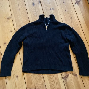 Svart stickad tröja med dragkedja från 157 - Svart stickad tröja från 157 med hög krage och dragkedja framtill. Tröjan har lång ärm och är ribbad i materialet, vilket ger en snygg och enkel look. Perfekt för kyligare dagar nu på våren. Storlek S men passar nog också M