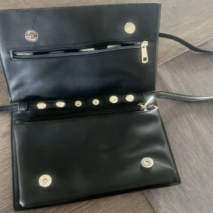 Svart axelväska med nitar - Snygg svart axelväska med silverfärgade nitar i stjärnmönster. Väskan har dragkedja och magnetknäppning samt en smal axelrem. Perfekt för att lyfta din outfit med en cool detalj.