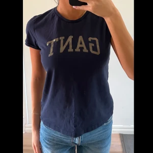 Mörkblå t-shirt från GANT - Mörkblå t-shirt från GANT med klassisk rund halsringning och tryckt logga framtill i vitt. T-shirten har en enkel och stilren design med korta ärmar och normal passform. Perfekt till jeans eller shorts.