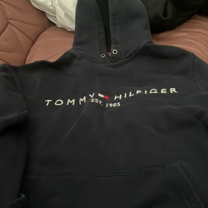 Mörkblå hoodie från Tommy Hilfiger - Säljer en mörkblå hoodie från Tommy Hilfiger med logga och texten 'EST. 1985' på bröstet. Tröjan har huva med dragsko och en stor magficka framtill. Perfekt för en avslappnad stil.