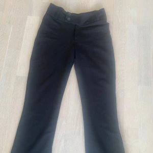 Svarta kostym byxor med knappar - Säljer ett par svarta byxor med  två dekorativa knappar framtill. Byxorna har en bootcut/rak  passform och är tillverkade i ett mjukt material. Uppsydda så passar 158 ish men ej klippta i så går att spretta upp lätt. Gina tricot Young
