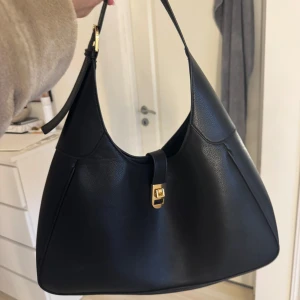 Svart axelväska med gulddetalj - Snygg svart axelväska i skinn med stilren design och guldfärgat spänne framtill. Väskan har en klassisk form och ett brett axelband som gör den bekväm att bära. Perfekt för att lyfta vilken outfit som helst.