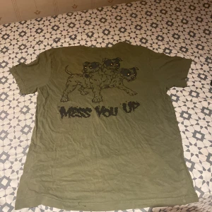 Olivgrön CrossFit t-shirt med tryck - Säljer en olivgrön t-shirt från CrossFit med tryck på både fram- och baksidan. På ryggen finns en stor illustration av en trehövdad hund och texten 'MESS YOU UP'. Perfekt för dig som gillar unika träningsplagg.
