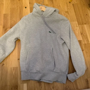 Grå hoodie från Lacoste - Säljer en klassisk grå hoodie från Lacoste med broderad logga på bröstet. Tröjan har huva med dragsko och en stor magficka. Perfekt för en avslappnad stil.