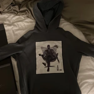 Mörkgrå hoodie från Limitato - Säljer en mörkgrå hoodie från Limitato med en stor grafisk print på framsidan som föreställer en person i kostym med hatt, cigarr och flaska. Hoodien har en klassisk passform och är tillverkad i mjukt material. Perfekt för dig som gillar unika tryck. Köpt på nk för ca 3000 kr. Skick 9/10. Box medföljer.
