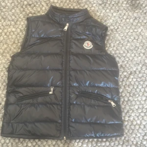 Svart dunväst från Moncler - Säljer en svart glansig dunväst från Moncler med hög krage och dragkedja framtill. Västen har två snedställda fickor med dragkedja och Moncler-logga på bröstet. Perfekt för lager-på-lager-stil.