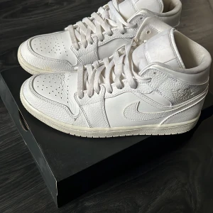 Nike Air Jordan 1 Mid White - Säljer ett par helvita Nike Air Jordan 1 Mid sneakers i skinn. Klassisk siluett med perforerad tå, snörning och ikoniska detaljer. Perfekta för dig som gillar stilrena och tidlösa sneakers. Kommer i originalkartong.