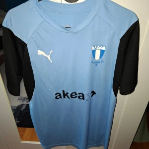 Malmö FF fotbollströja Puma - Ljusblå Malmö FF matchtröja från Puma med svarta ärmar och vita loggor. Tryck med sponsor 'akea' på framsidan och 'MOBILL' på ryggen. Klassisk klubbsköld på bröstet. Perfekt för fotbollsfans!