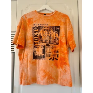 Orange t-shirt med Tokyo-tryck - Cool orange t-shirt med stort svartvitt Tokyo-tryck framtill och japanska tecken. Oversized passform och korta ärmar, perfekt för en avslappnad streetstyle-look.