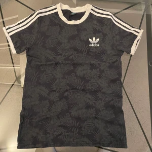 Svart mönstrad t-shirt från Adidas - Snygg svart t-shirt från Adidas med diskret bladmönster och vita detaljer. Klassiska tre ränder på axlarna och Adidas-logga på bröstet. Rund halsringning och korta ärmar. Perfekt för en sportig och avslappnad stil.