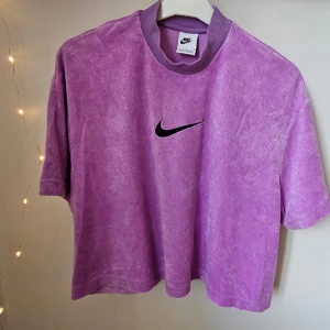 Lila t-shirt i frotté från Nike - Cropped t-shirt från Nike. Lila frotté med svart broderad swoosh-logga på bröstet. T-shirten har korta ärmar och hög krage. Tillverkad i en mjuk blandning av bomull, polyester och viskos. Perfekt för en avslappnad och sportig stil.