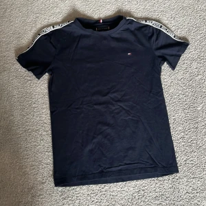 Mörkblå t-shirt från Tommy Hilfiger - Snygg mörkblå t-shirt från Tommy Hilfiger med logoband på axlarna och liten broderad logga på bröstet. Klassisk passform och rund halsringning. Perfekt för en stilren och sportig look.