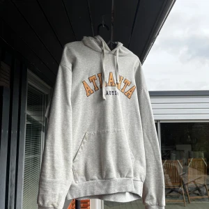 Grå hoodie  - Säljer en grå hoodie med huva och känguruficka framtill. På bröstet står det ATLANTA i stora oranga bokstäver med svart kontur. Tröjan har normal passform och är perfekt för en avslappnad stil.