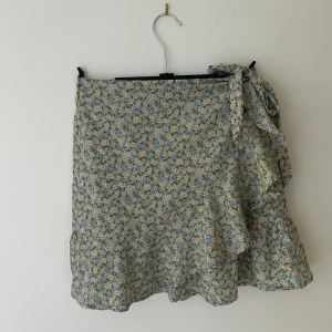 Blommig omlottkjol med volang - Supersöt kortkjol med blått och vitt blommönster. Kjolen har omlottmodell med knyt i sidan och volang nertill. Perfekt för sommarens alla tillfällen!