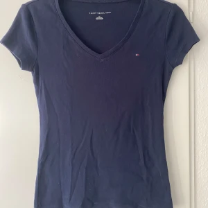 Mörkblå t-shirt från Tommy Hilfiger - Mörkblå t-shirt från Tommy Hilfiger med v-ringning och liten logga på bröstet. T-shirten har korta ärmar och är tillverkad i mjuk bomull. Perfekt till vardags och enkel att matcha med olika outfits.