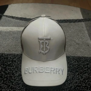 Burberry keps - Säljer en vit keps från Burberry med broderad logga framtill och texten 'BURBERRY' på skärmen. Sidorna och baksidan har det klassiska rutiga Burberry-mönstret. Justerbar rem baktill. Perfekt accessoar för en trendig look. Priset ej hugget i sten