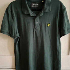 Mörkgrön pikétröja från Lyle & Scott - Snygg mörkgrön pikétröja från Lyle & Scott med klassisk krage och knappslå framtill. Tröjan har korta ärmar och en gul broderad logga på bröstet. Perfekt för en stilren och avslappnad look. Storlek L men tycker den är mer som M