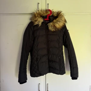 Svart jacka med fuskpäls - Svart pufferjacka med huva och fuskpälsdetalj. Jackan har dragkedja framtill och två fickor med dragkedja. Perfekt för kyligare dagar och har en klassisk, stilren look. Storlek S. Kan gå ner i pris vid snabb affär 