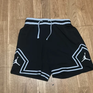Svarta Jordan shorts - Snygga svarta shorts från Jordan med vit snörning och ljusgrå detaljer längs midjan och benen. Ikonisk Jumpman-logga på båda benen. Perfekta för sport eller chill dagar.