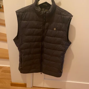Mörkgrön dunväst från Morris - Snygg mörkgrön dunväst från Morris med quiltad design och dragkedja framtill. Västen har en liten broderad logga på bröstet och hög krage. Perfekt att bära över en tröja för extra värme. Nypris 2299kr